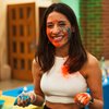 A smiling young woman with paint smeared on her face and hands holds blue and orange powder in a colorful indoor setting.