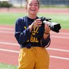 A smiling young woman stands on a running track holding a large camera with a telephoto lens.
