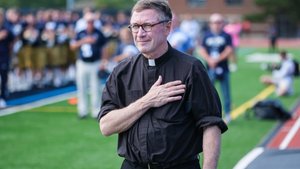 A man wearing a black clerical shirt with a white collar stands on a sports field with his right hand over his heart.