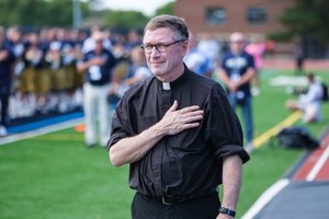 A man wearing a black clerical shirt with a white collar stands on a sports field with his right hand over his heart.