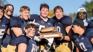 A group of football players in navy and gold uniforms smile and hold a wooden trophy with a football on top under a clear.