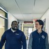 Two men wearing dark blue John Carroll University clothing walk and smile at each other in a hallway.