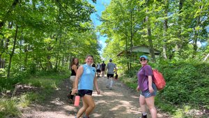 A group of people walk up a dirt path surrounded by green trees under a blue sky, with two smiling women facing the camera.
