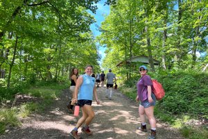 A group of people walk up a dirt path surrounded by green trees under a blue sky, with two smiling women facing the camera.