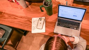 A person with braided hair sits at a wooden table using a laptop with a notebook, phone, and drink nearby.