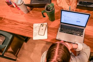 A person with braided hair sits at a wooden table using a laptop with a notebook, phone, and drink nearby.
