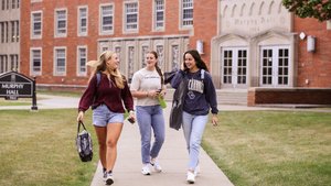 Three young women walk and talk on a sidewalk in front of a brick building labeled Murphy Hall.