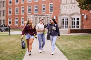 Three young women walk and talk on a sidewalk in front of a brick building labeled Murphy Hall.