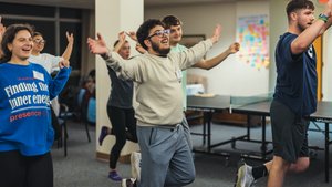 A group of young people indoors stand on one leg with arms raised, smiling and balancing near a ping pong table.