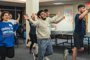 A group of young people indoors stand on one leg with arms raised, smiling and balancing near a ping pong table.