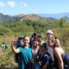 A group of six young people pose closely together smiling for a selfie on a sunny hillside with mountains in the background.