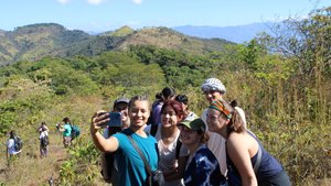 A group of six young people pose closely together smiling for a selfie on a sunny hillside with mountains in the background.
