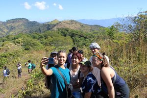 A group of six young people pose closely together smiling for a selfie on a sunny hillside with mountains in the background.