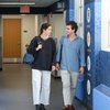 Two young people walk side by side in a hallway, smiling and carrying backpacks.