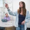 A young woman writes mathematical equations on a glass board while another woman watches from a table with a laptop.