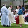 Two young women hand a golden bowl to a priest outdoors while people seated on white chairs watch.