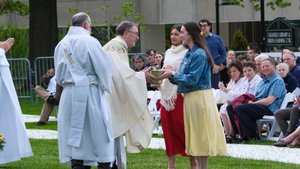 Two young women hand a golden bowl to a priest outdoors while people seated on white chairs watch.
