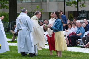 Two young women hand a golden bowl to a priest outdoors while people seated on white chairs watch.