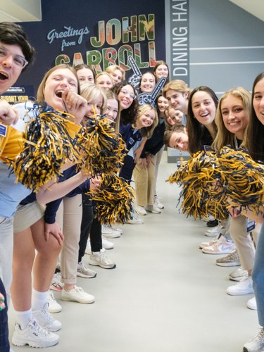 A group of young people cheer and smile while holding blue and gold pom-poms in a hallway with a John Carroll sign.