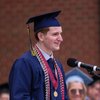 A young man in a blue graduation gown and cap speaks at a microphone with a blurred audience behind him.