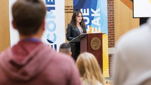 A woman with curly hair speaks at a podium in front of banners while an audience listens indoors.