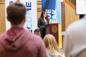 A woman with curly hair speaks at a podium in front of banners while an audience listens indoors.