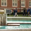 A group of seven people walk along a sidewalk beside a brick building near a fountain with water flowing.