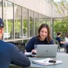 Two students sit at a table outside a building, one smiling at a laptop while the other faces away wearing a cap.