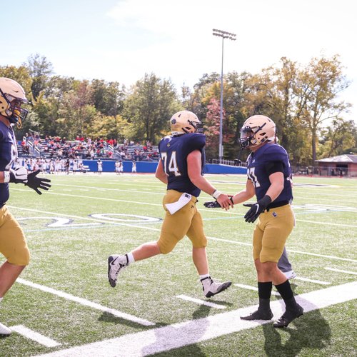 Three football players in navy and gold uniforms are on a sunlit field, with two shaking hands near the sideline.