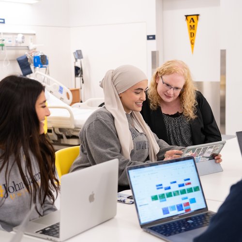 Four students sit around a table with laptops, smiling and looking at a tablet in a room with hospital beds.