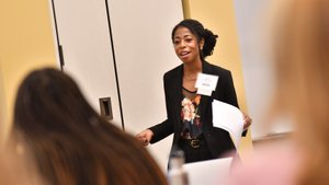 A woman wearing a black blazer and floral top is speaking and holding papers in front of an audience.