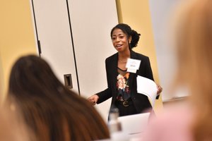 A woman wearing a black blazer and floral top is speaking and holding papers in front of an audience.