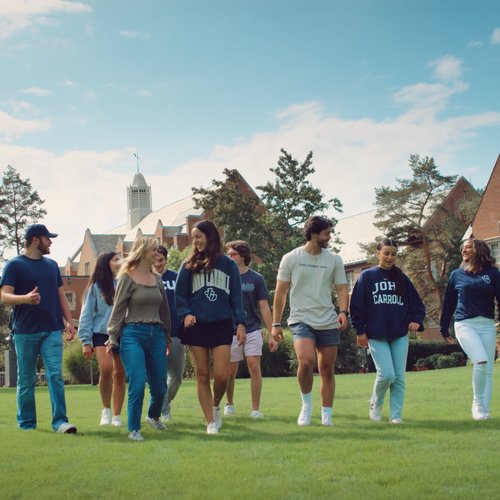 A group of eight young adults walk and talk together on a green lawn in front of large brick buildings under a partly cloudy.