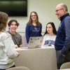 A group of four people, three young adults and one older man, are gathered around a laptop in a classroom.
