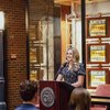 A young woman with long blonde hair smiles while speaking at a podium in front of an audience indoors.