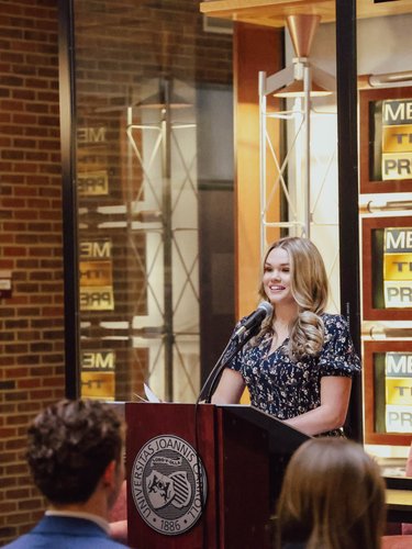 A young woman with long blonde hair smiles while speaking at a podium in front of an audience indoors.