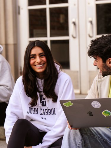 Three young adults sit on steps outside a building, two holding laptops and smiling at each other.