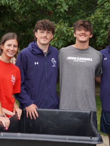 Six young adults stand outdoors behind a large black plastic container, smiling at the camera under green trees.