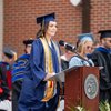 A female graduate in a blue cap and gown speaks at a podium during an outdoor ceremony with faculty standing behind her.