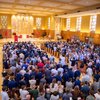 A large group of people stands inside a church facing the altar with stained glass windows and religious statues.