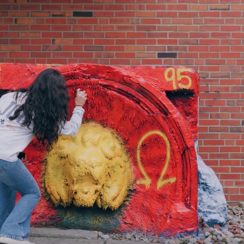 A woman spray paints a red and yellow concrete sculpture while a man watches against a brick wall background.