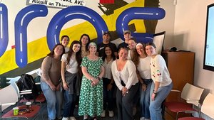 A group of eleven people stand closely together smiling in a room with colorful wall art and empty chairs around them.