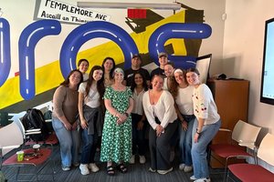 A group of eleven people stand closely together smiling in a room with colorful wall art and empty chairs around them.