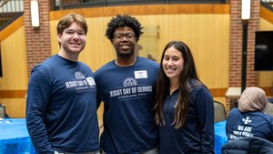 Three young adults wearing blue shirts smile and pose together inside a room with brick walls and blue-covered tables.