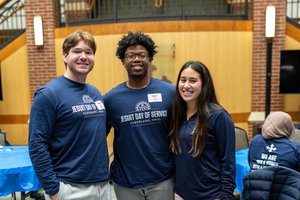Three young adults wearing blue shirts smile and pose together inside a room with brick walls and blue-covered tables.