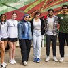 Six young adults stand arm in arm smiling in front of a colorful mural on a brick wall, one holding a shovel.
