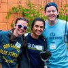 Three young people smile at the camera while holding gardening tools against an orange wall with vines.