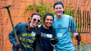 Three young people smile at the camera while holding gardening tools against an orange wall with vines.