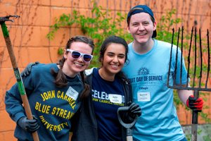 Three young people smile at the camera while holding gardening tools against an orange wall with vines.