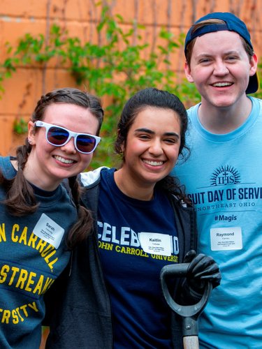 Three young people smile at the camera while holding gardening tools against an orange wall with vines.